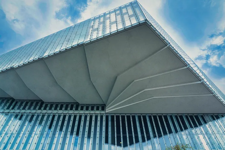 Underside of a modern glass-clad building with folded concrete soffit against a blue sky